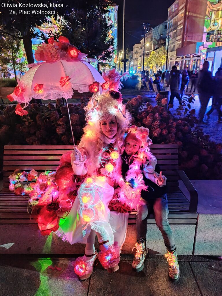 Oliwia z 2 a - dziewczynka siedzi na ławce przytulona do pięknej, kolorowej pani z parasolką i światełkami