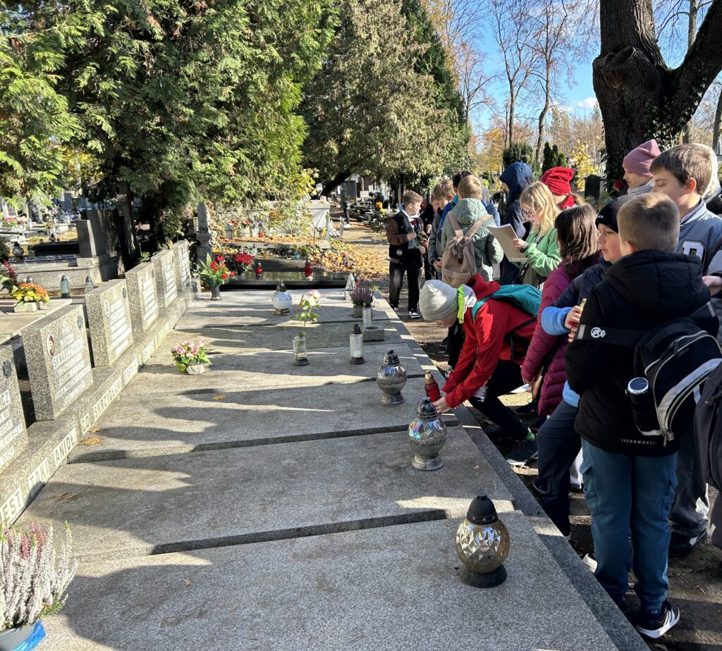 dzieci stoją przy grobach harcerek, jeden z chłopców kładzie zapalony znicz