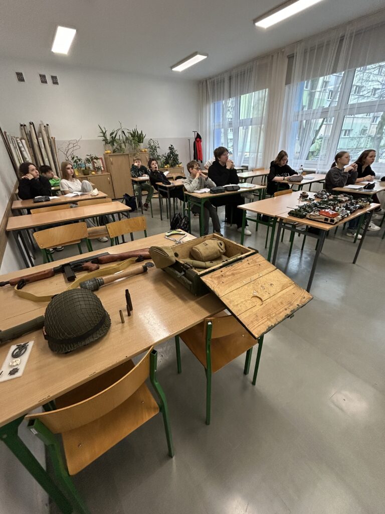 klasa, w niej uczniowie na pierwszych ławkach widać eksponaty - sprzęt wojskowy, np. hełm,  oraz pojazdy wojskowe z klocków