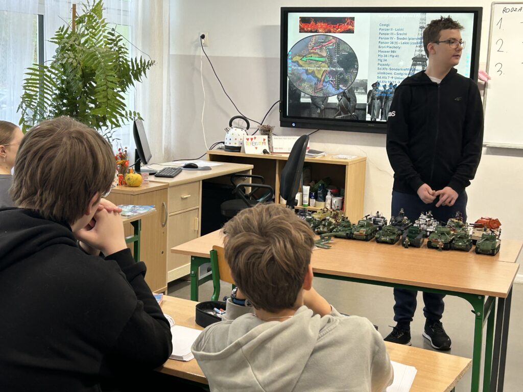 chłopiec w klasie opowiada o pojazdach wojskowych, które stoją na ławce, za nim widoczny tablica multimedialna z prezentacją o uzbrojeniu lądowym w 2 wojnie św. tyłem do aparatu w ławce siedzący dwaj chłopcy