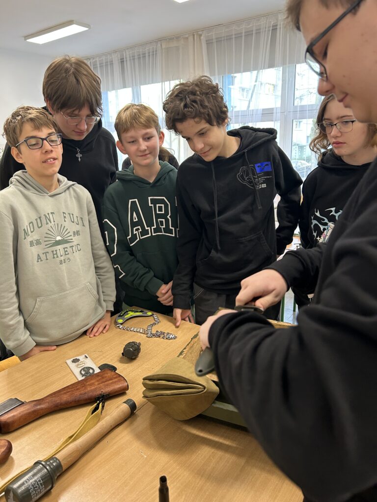 na dwóch ławkach położony sprzęt wojskowy, wokół ławek stoją uczniowie i patrzą na prezentowane przedmioty