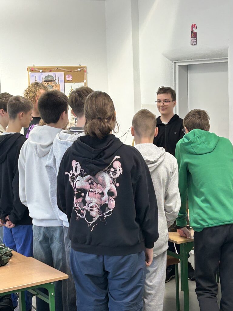 uczniowie stoją tyłem do aparatu przed nimi chłopiec opowiada o swojej kolekcji sprzętu wojskowego