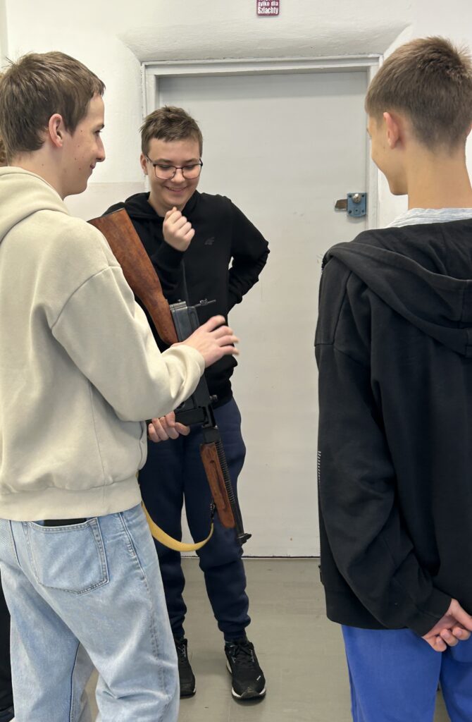 trzej chłopcy stoją i rozmawiają, jeden z nich trzyma w rękach karabin skierowany do dołu