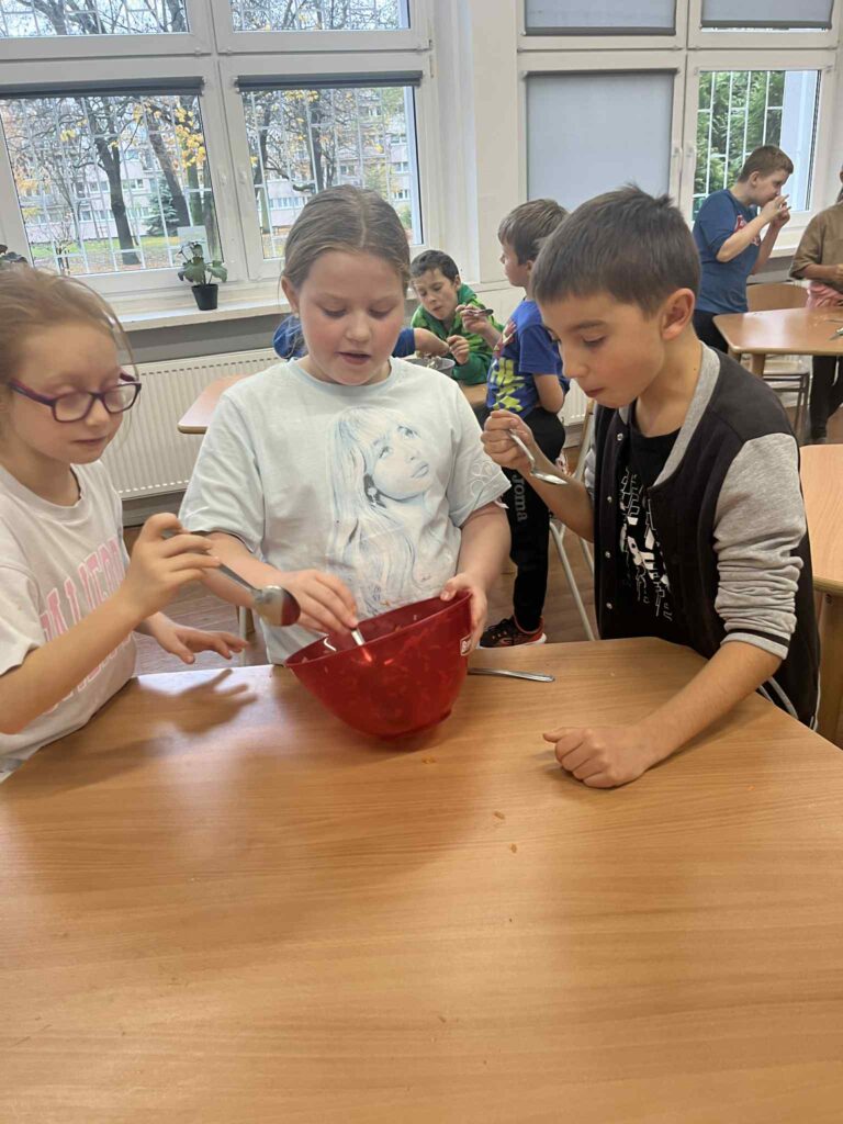 Troje uczniów miesza łyżkami w czerwonej misce przy stoliku.