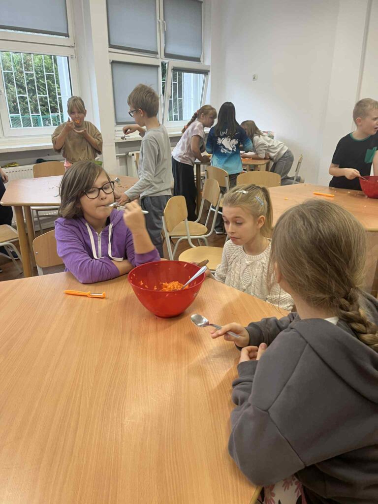 Na pierwszym planie trzy dziewczynki próbują łyżeczkami surówkę z czerwonej miseczki. W tle pozostali uczniowie przy stolikach.