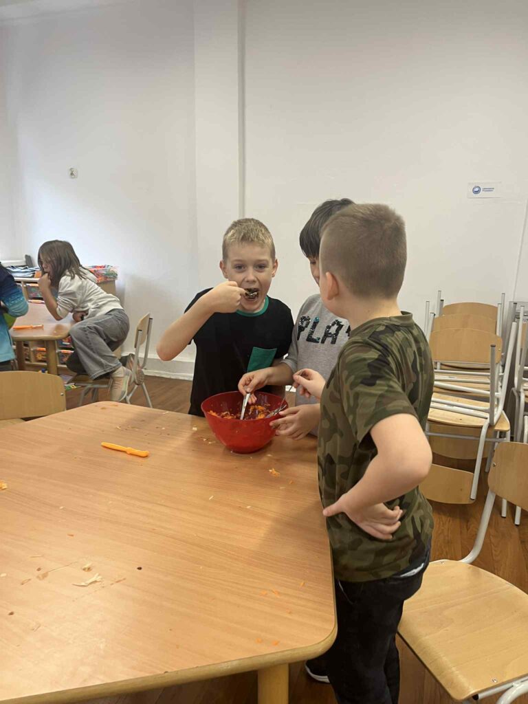 Trzech chłopców próbuje łyżeczkami marchewkowej surówki z czerwonej miseczki