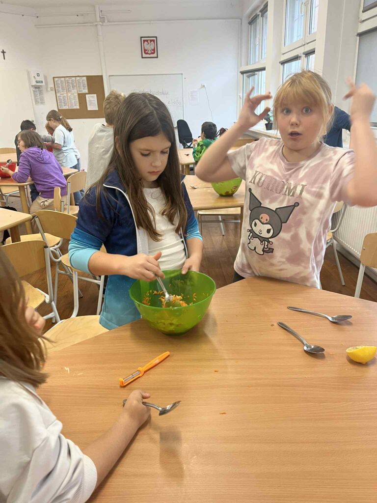 Dwie uczennice mieszają łyżkami marchewkową surówkę w zielonej miseczce.