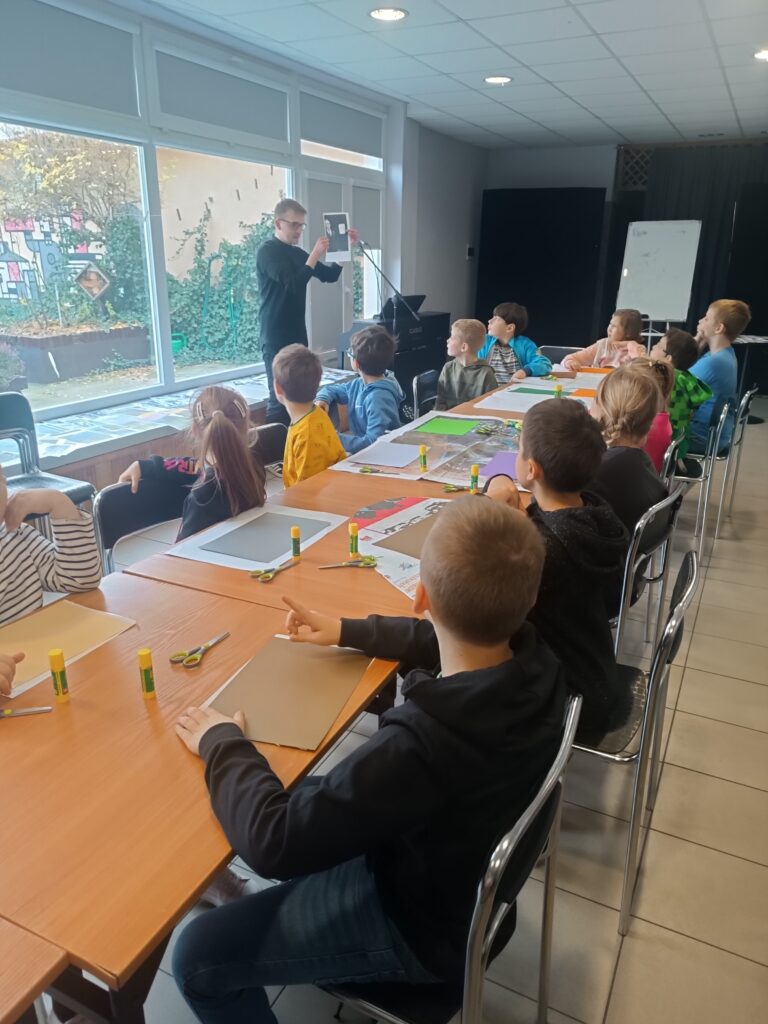 Uczniowie siedzą przy długim stole z przygotowanymi przed sobą kartkami i słuchają mówiącego do nich mężczyzny