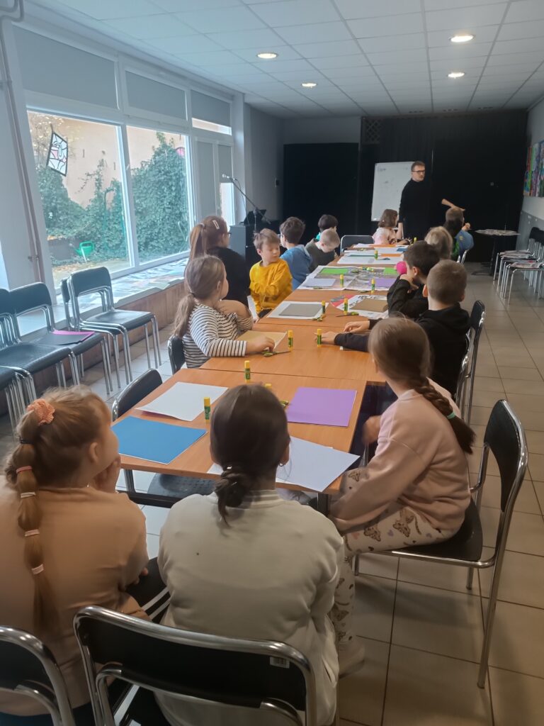 Uczniowie siedzą przy długim stole z przygotowanymi przed sobą kartkami i słuchają mówiącego do nich mężczyzny