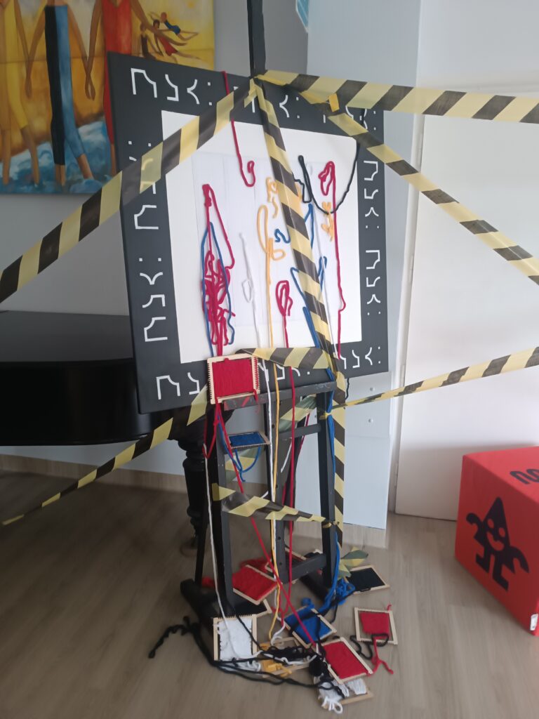 Instalacja artystyczna złożona z włóczek w różnym kolorze, krosen, taśmy ostrzegawczej czarno-żółtej. Wszystko stoi na stojaku. 