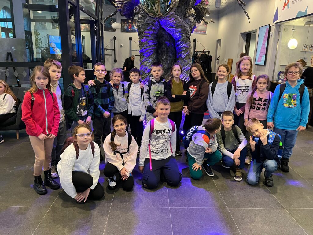 Uczniowie pozują do grupowego zdjęcia w korytarzu teatru, pod dekoracją przypominającą nogi ogromnego człowieka