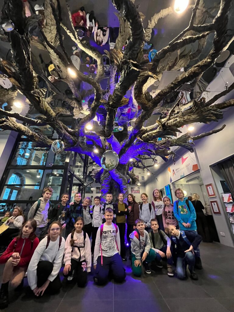 Uczniowie pozują do grupowego zdjęcia w korytarzu teatru, pod dekoracją przypominającą ogromne drzewo z wiszącymi na gołych gałęziach lustrzanymi kulami