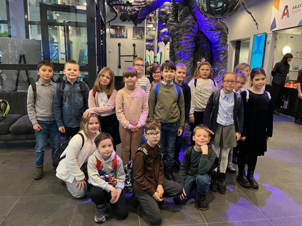 Uczniowie pozują do grupowego zdjęcia w korytarzu teatru, pod dekoracją przypominającą nogi ogromnego człowieka.