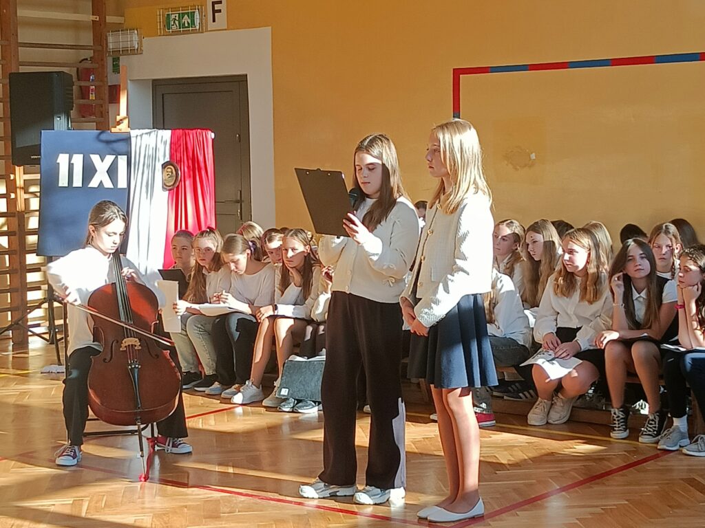 uczennica gra na kontrabasie, dwie uczennice stoją obok, za nimi siedzi chór. 