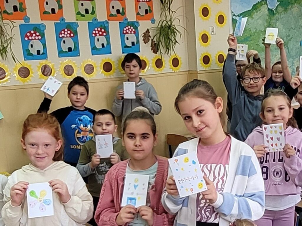 uczniowie i uczennice trzymają karteczki  będące laurkami, uśmiechają się. W tle kolorowa klasa przystrojona kolorowymi rysunkami.