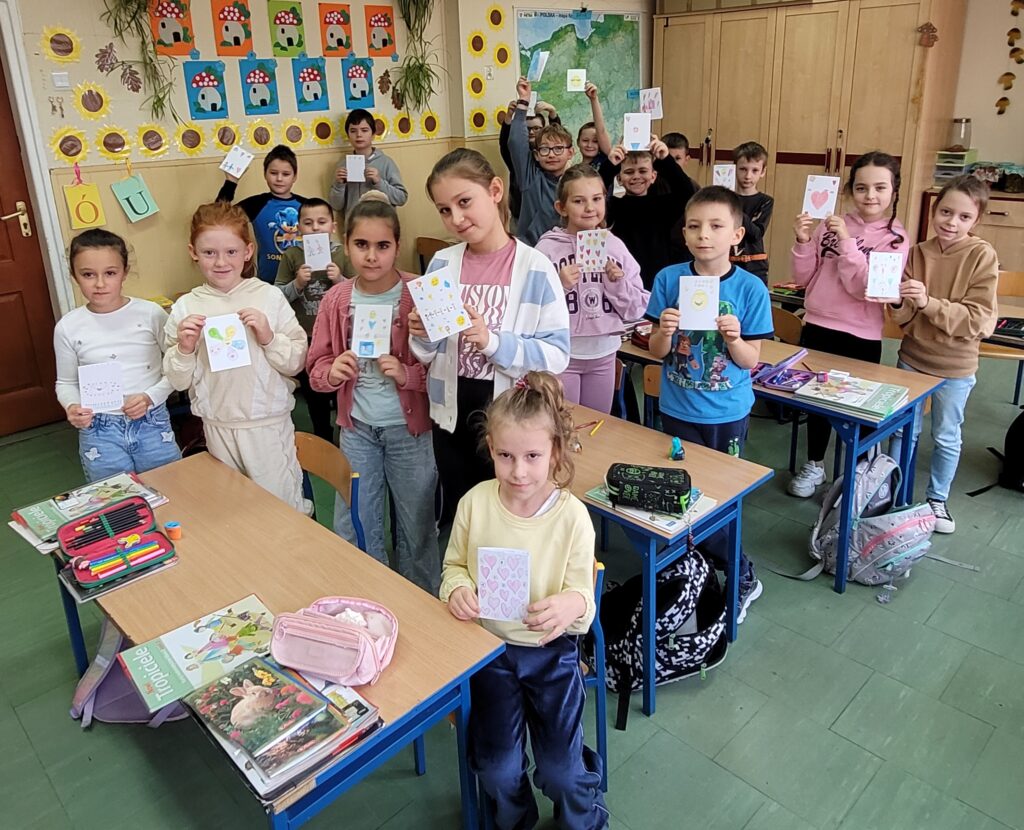 uczniowie i uczennice trzymają karteczki  będące laurkami, uśmiechają się. W tle kolorowa klasa przystrojona kolorowymi rysunkami.