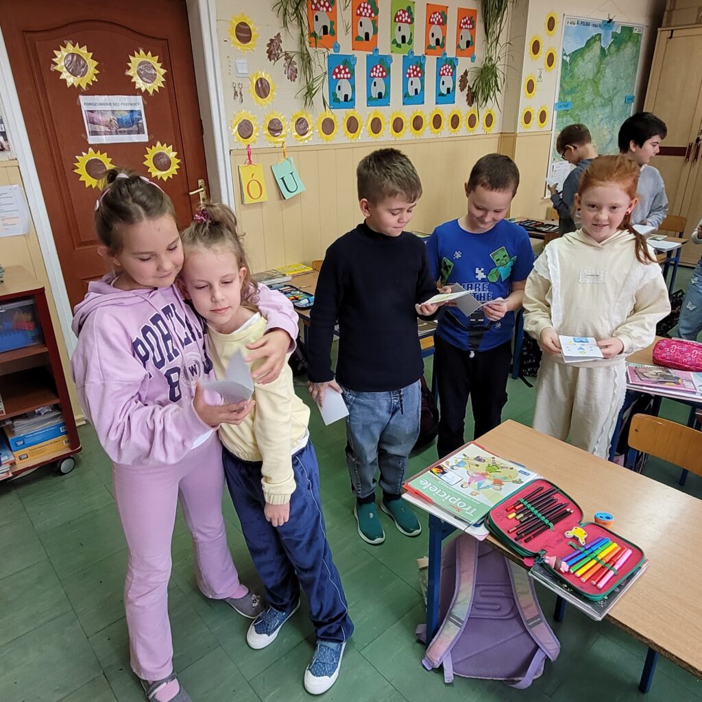 uczniowie i uczennice trzymają karteczki  będące laurkami, uśmiechają się. W tle kolorowa klasa przystrojona kolorowymi rysunkami.