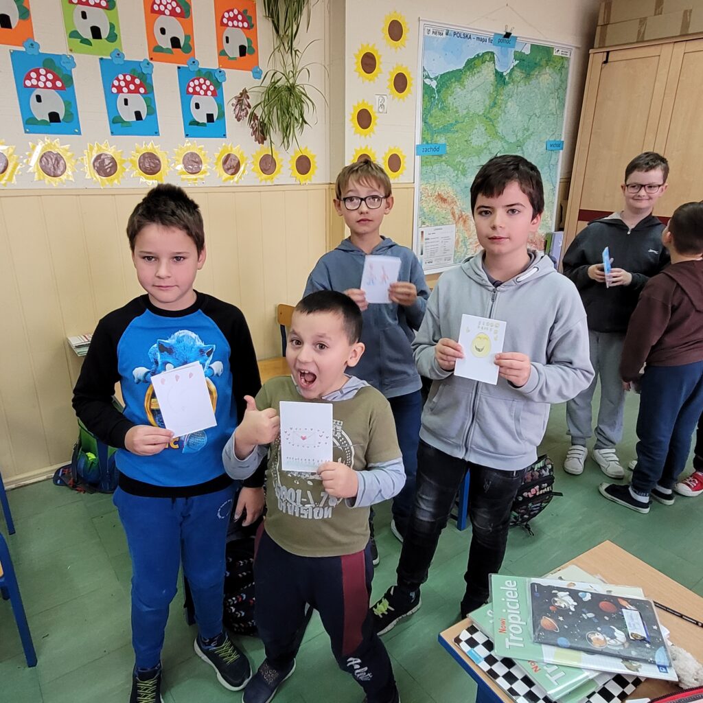 uczniowie trzymają karteczki  będące laurkami, uśmiechają się. W tle kolorowa klasa przystrojona kolorowymi rysunkami.