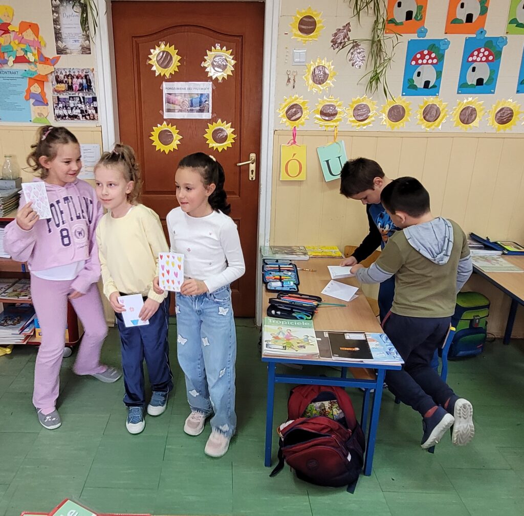 uczniowie i uczennice trzymają karteczki  będące laurkami, uśmiechają się. W tle kolorowa klasa przystrojona kolorowymi rysunkami.