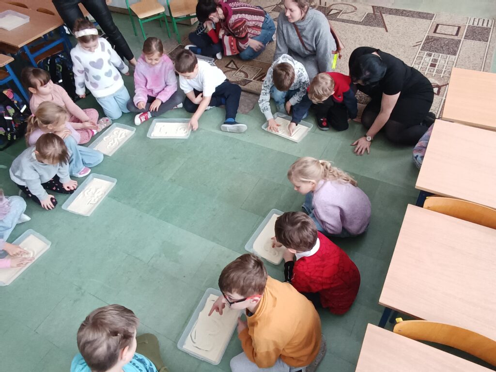 dzieci rysują palcem po piasku, piasek w pojemnikach plastikowych, zdjęcie z góry, widać wiele osób 