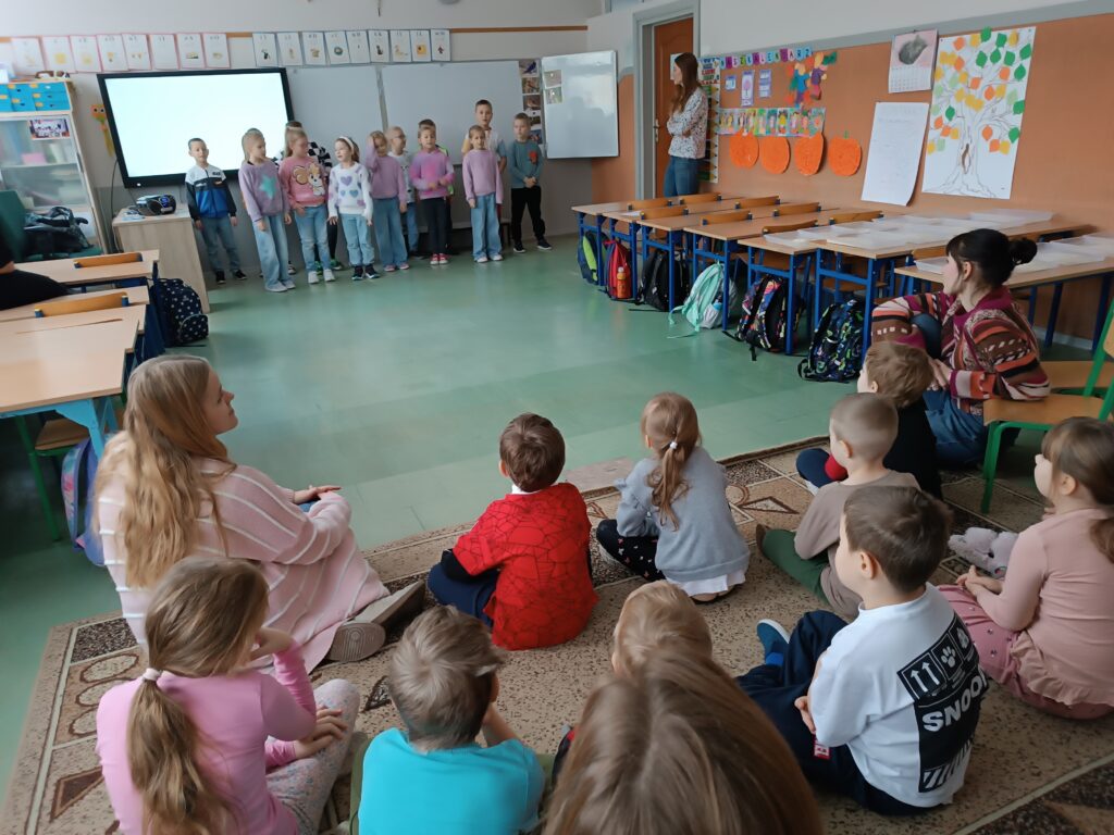 z przodu dzieci odwrócone plecami, siedzą na dywanie, z tyłu dzieci stoją i mówią coś do dzieci siedzących