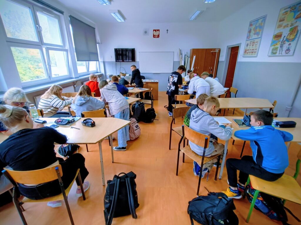 zdjęcie przedstawia uczniów siedzących w ławkach i wykonujących zadania.