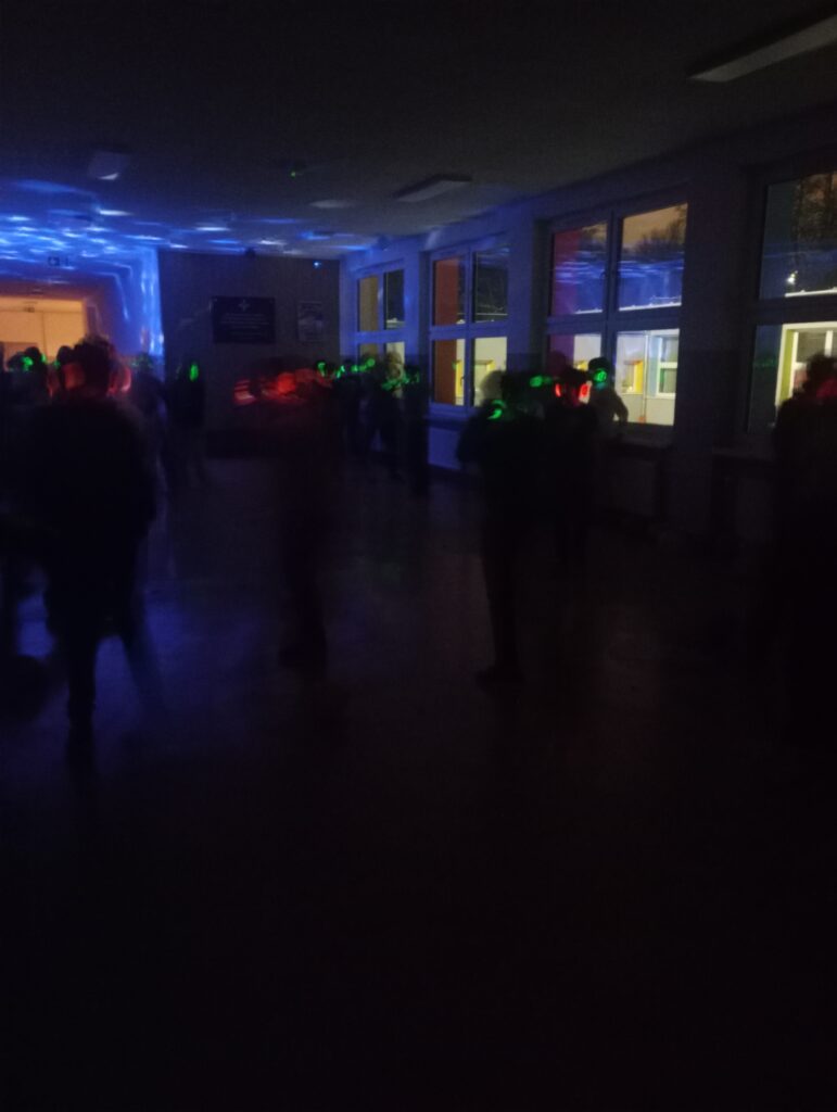 Dyskoteka. Uczniowie w świecących słuchawkach na głowach stoją w korytarzu w grupkach, jest półmrok a na ścianach kolorowe refleksy zielono, niebiesko, czerwone
