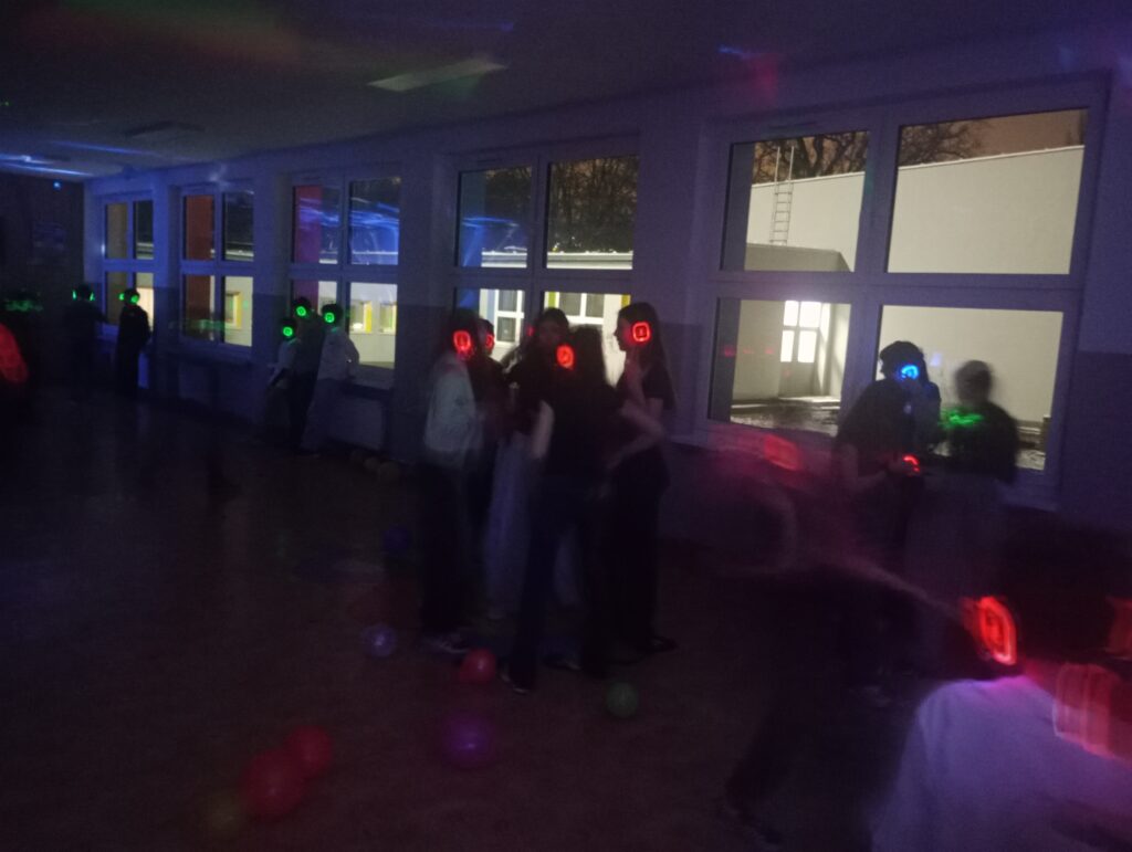 Uczniowie w świecących słuchawkach na głowach stoją w korytarzu w grupkach, jest półmrok a na ścianach kolorowe refleksy zielono, niebiesko, czerwone