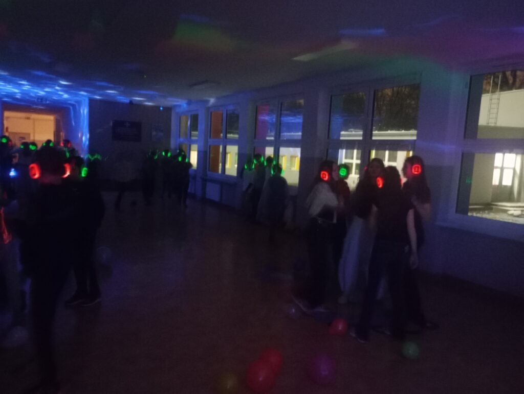 Dyskoteka. Uczniowie w świecących słuchawkach na głowach stoją w korytarzu w grupkach, jest półmrok a na ścianach kolorowe refleksy zielono, niebiesko, czerwone