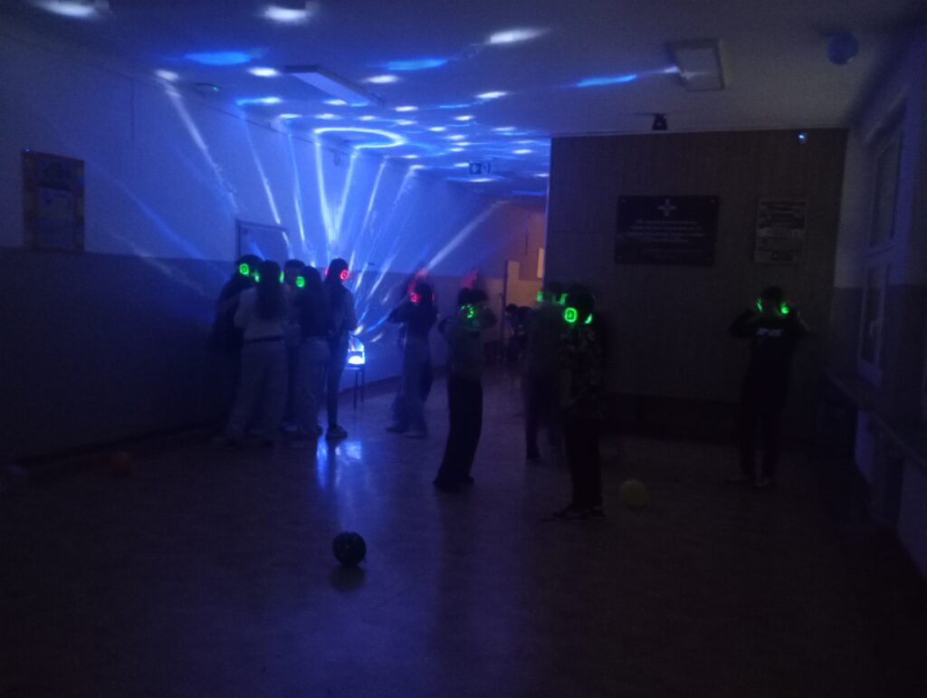 Dyskoteka. Uczniowie w świecących słuchawkach na głowach stoją w korytarzu w grupkach, jest półmrok a na ścianach kolorowe refleksy zielono, niebiesko, czerwone