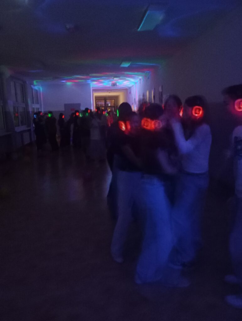 Dyskoteka. Uczniowie w świecących słuchawkach na głowach stoją w korytarzu w grupkach, jest półmrok a na ścianach kolorowe refleksy zielono, niebiesko, czerwone