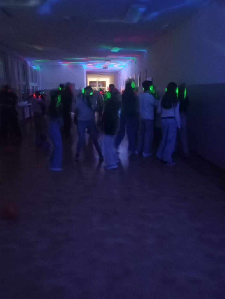 Dyskoteka. Uczniowie w świecących słuchawkach na głowach stoją w korytarzu w grupkach, jest półmrok a na ścianach kolorowe refleksy zielono, niebiesko, czerwone