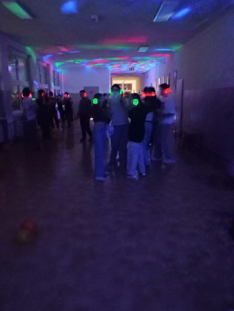 Dyskoteka. Uczniowie w świecących słuchawkach na głowach stoją w korytarzu w grupkach, jest półmrok a na ścianach kolorowe refleksy zielono, niebiesko, czerwone