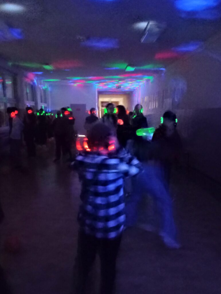 Dyskoteka. Uczniowie w świecących słuchawkach na głowach stoją w korytarzu w grupkach, jest półmrok a na ścianach kolorowe refleksy zielono, niebiesko, czerwone