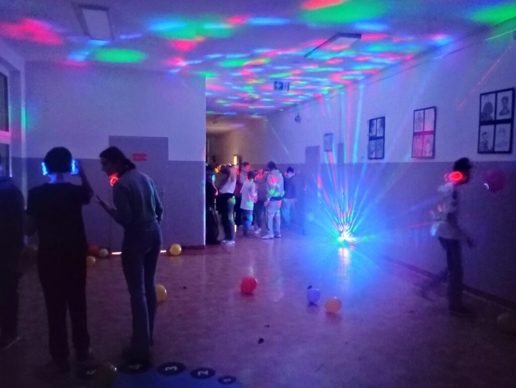 Dyskoteka. Uczniowie w świecących słuchawkach na głowach stoją w korytarzu w grupkach, jest półmrok a na ścianach kolorowe refleksy zielono, niebiesko, czerwone