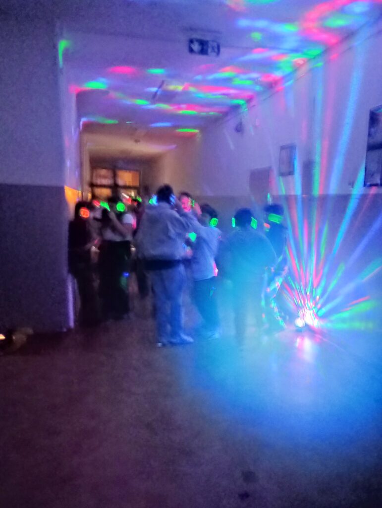 Dyskoteka. Uczniowie w świecących słuchawkach na głowach stoją w korytarzu w grupkach, jest półmrok a na ścianach kolorowe refleksy zielono, niebiesko, czerwone