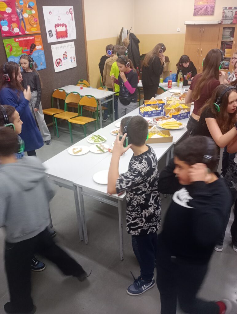 W pomieszczeniu ustawiony na środku stół z jedzeniem, dookoła dzieci w słuchawkach jedzą i piją soki