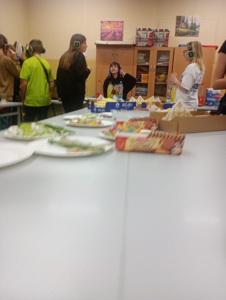 W pomieszczeniu ustawiony na środku stół z jedzeniem, dookoła dzieci w słuchawkach jedzą i piją soki