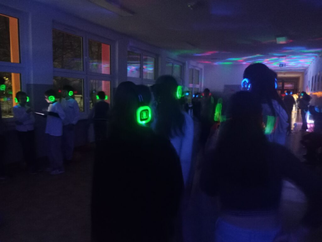 Dyskoteka. Uczniowie w świecących słuchawkach na głowach stoją w korytarzu w grupkach, jest półmrok a na ścianach kolorowe refleksy zielono, niebiesko, czerwone