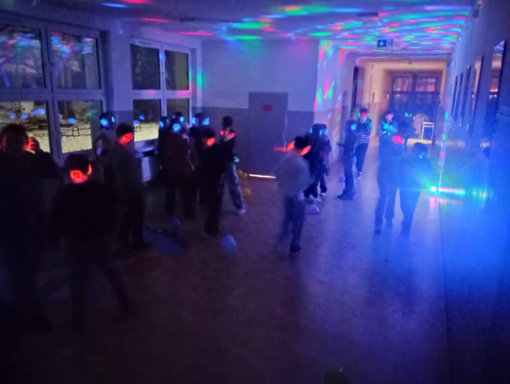 Dyskoteka. Uczniowie w świecących słuchawkach na głowach stoją w korytarzu w grupkach, jest półmrok a na ścianach kolorowe refleksy zielono, niebiesko, czerwone