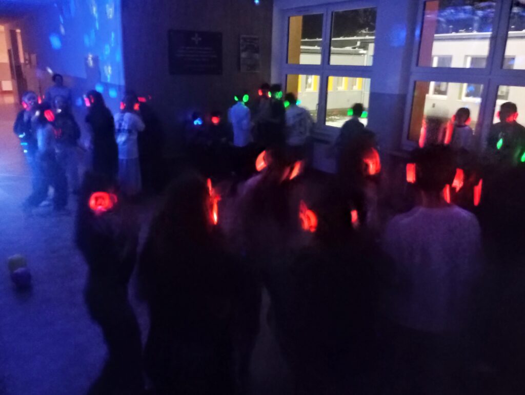 Dyskoteka. Uczniowie w świecących słuchawkach na głowach stoją w korytarzu w grupkach, jest półmrok a na ścianach kolorowe refleksy zielono, niebiesko, czerwone