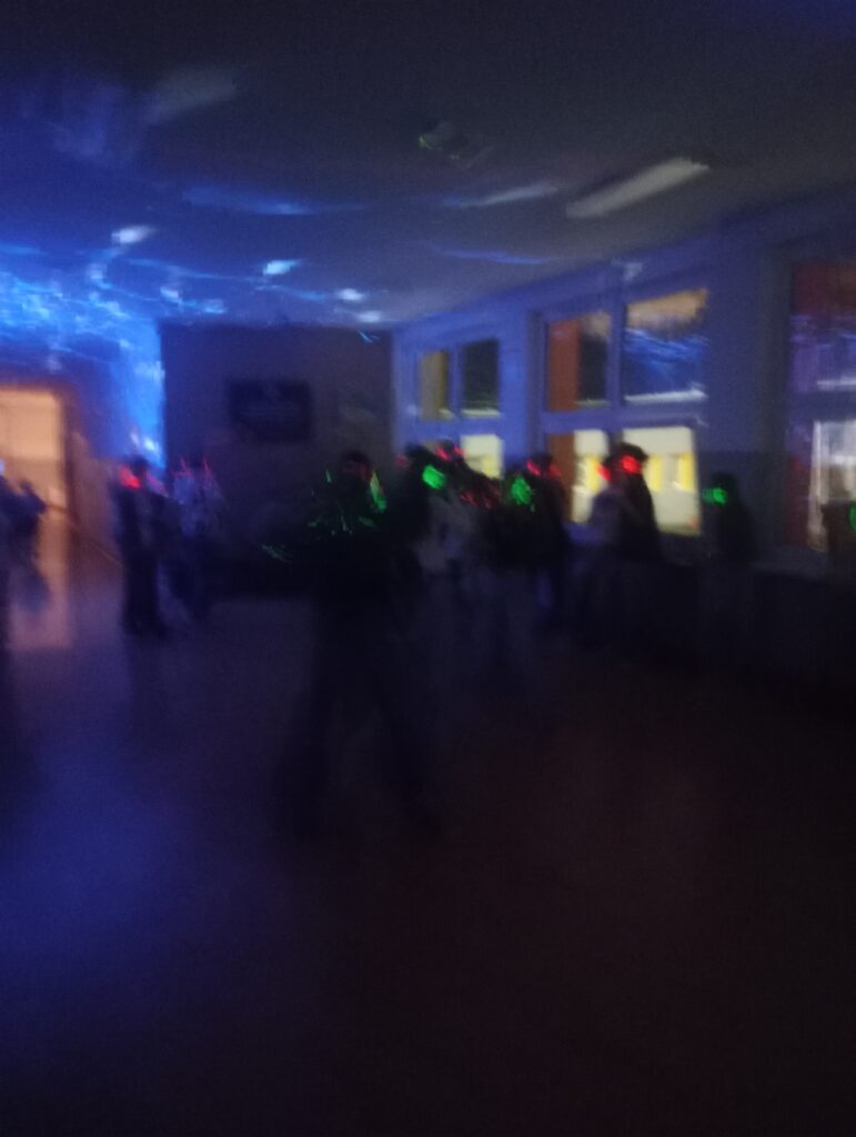 Dyskoteka. Uczniowie w świecących słuchawkach na głowach stoją w korytarzu w grupkach, jest półmrok a na ścianach kolorowe refleksy zielono, niebiesko, czerwone