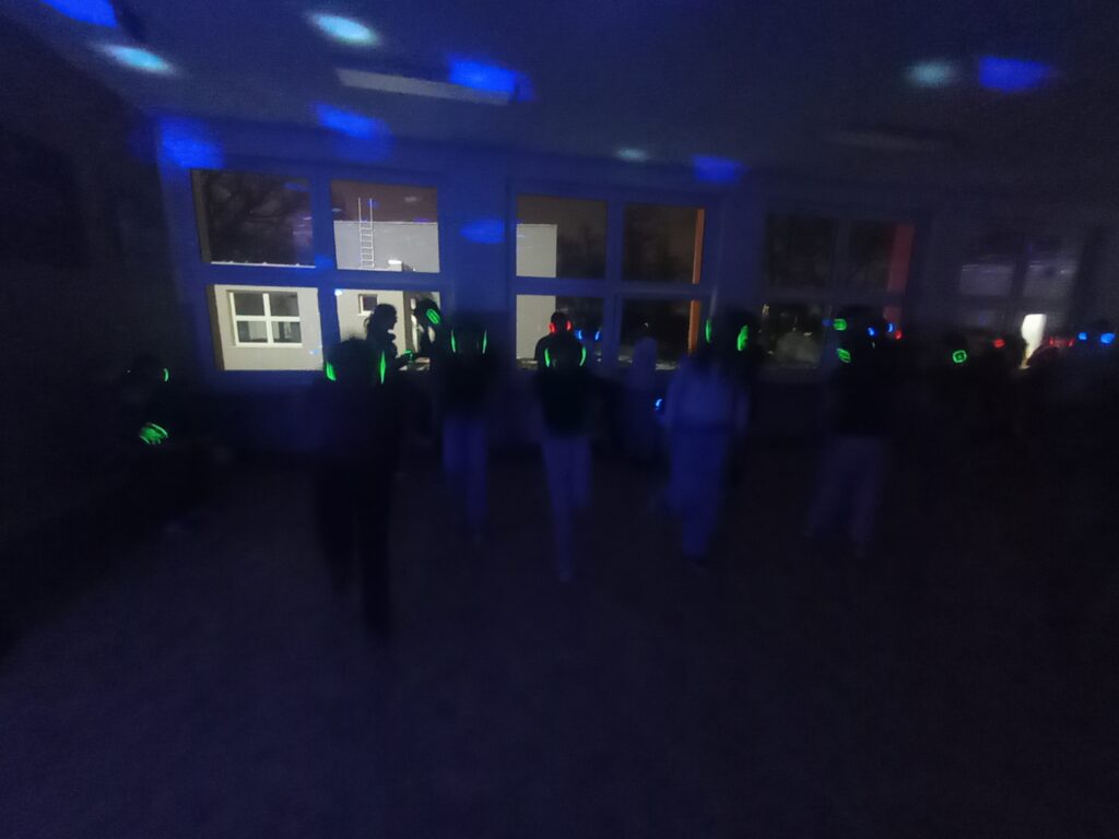 Dyskoteka. Uczniowie w świecących słuchawkach na głowach stoją w korytarzu w grupkach, jest półmrok a na ścianach kolorowe refleksy zielono, niebiesko, czerwone