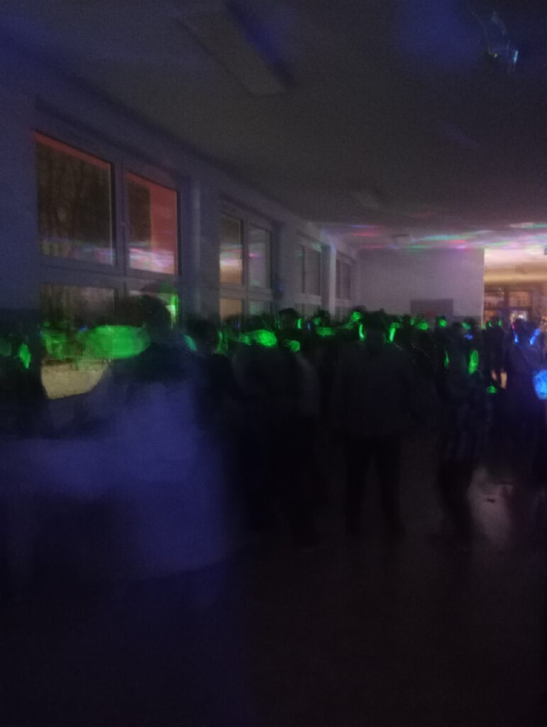 Dyskoteka. Uczniowie w świecących słuchawkach na głowach stoją w korytarzu w grupkach, jest półmrok a na ścianach kolorowe refleksy zielono, niebiesko, czerwone