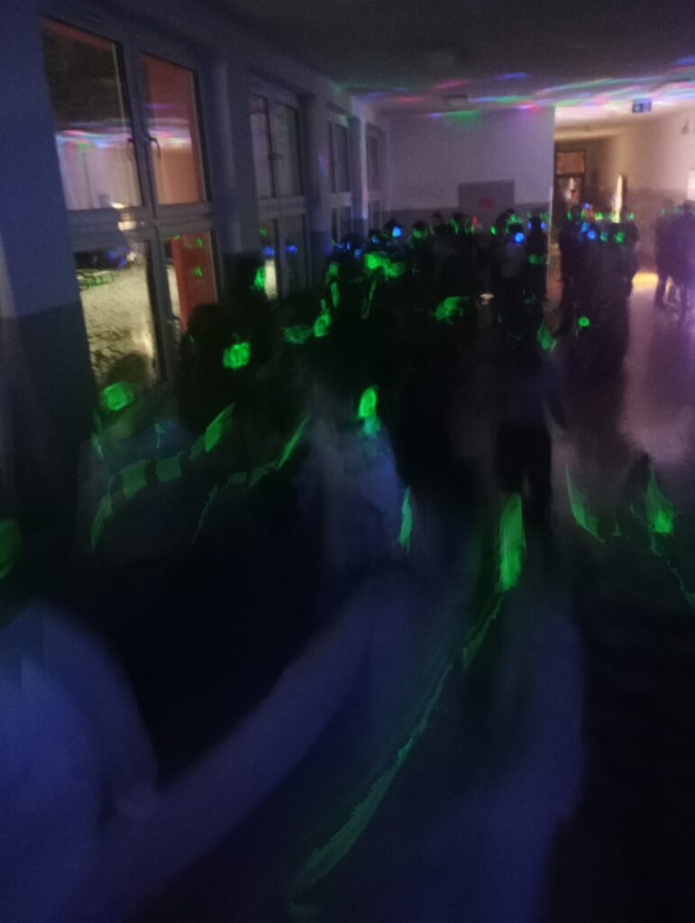 Dyskoteka. Uczniowie w świecących słuchawkach na głowach stoją w korytarzu w grupkach, jest półmrok a na ścianach kolorowe refleksy zielono, niebiesko, czerwone