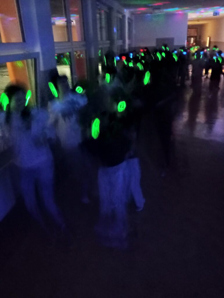 Dyskoteka. Uczniowie w świecących słuchawkach na głowach stoją w korytarzu w grupkach, jest półmrok a na ścianach kolorowe refleksy zielono, niebiesko, czerwone