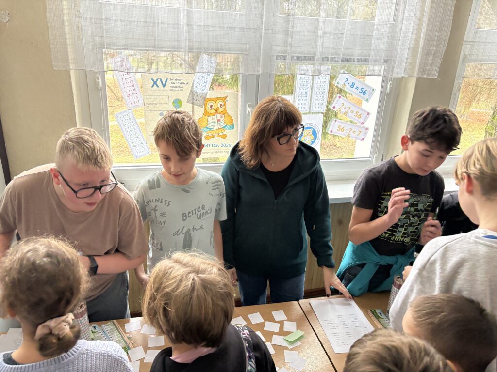 widzimy kolejkę uczniów czekających i odpowiadających z tabliczki mnożenia przed uczniowskimi komisjami, na ławkach widać  linijki, losy z pytaniami i ołówki