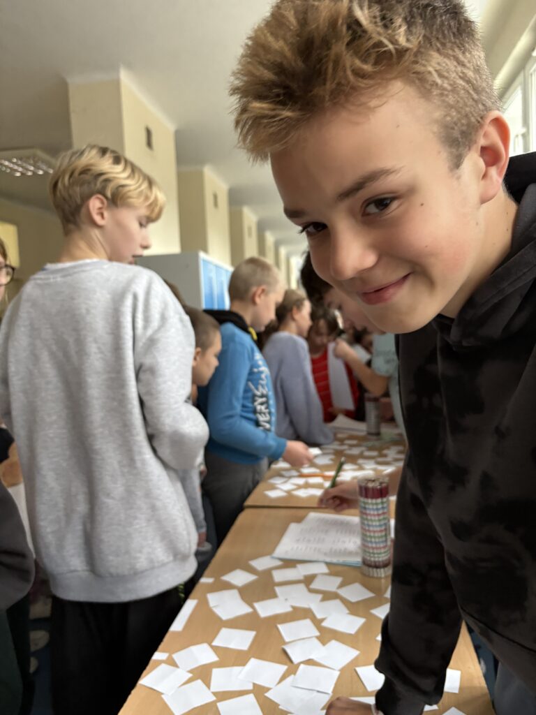 widzimy kolejkę uczniów czekających i odpowiadających z tabliczki mnożenia przed uczniowskimi komisjami, na ławkach widać  linijki, losy z pytaniami i ołówki