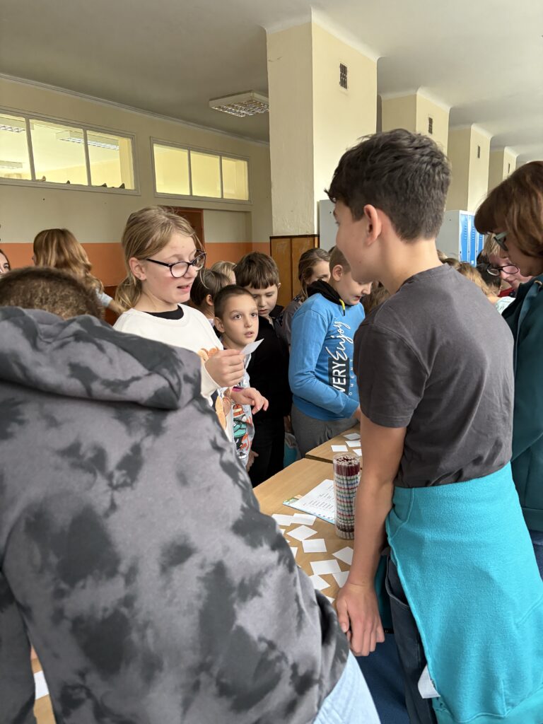 widzimy kolejkę uczniów czekających i odpowiadających z tabliczki mnożenia przed uczniowskimi komisjami, na ławkach widać  linijki, losy z pytaniami i ołówki