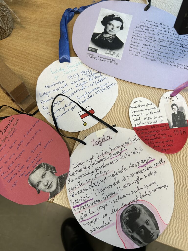 na ławce położone są zawieszki kartonowe ze wstążeczkami,  z informacjami o uczestniczkach powstania warszawskiego. Każda zawieszka poświęcona jest jednej kobiecie. Na dwóch są obok informacji także zdjęcia